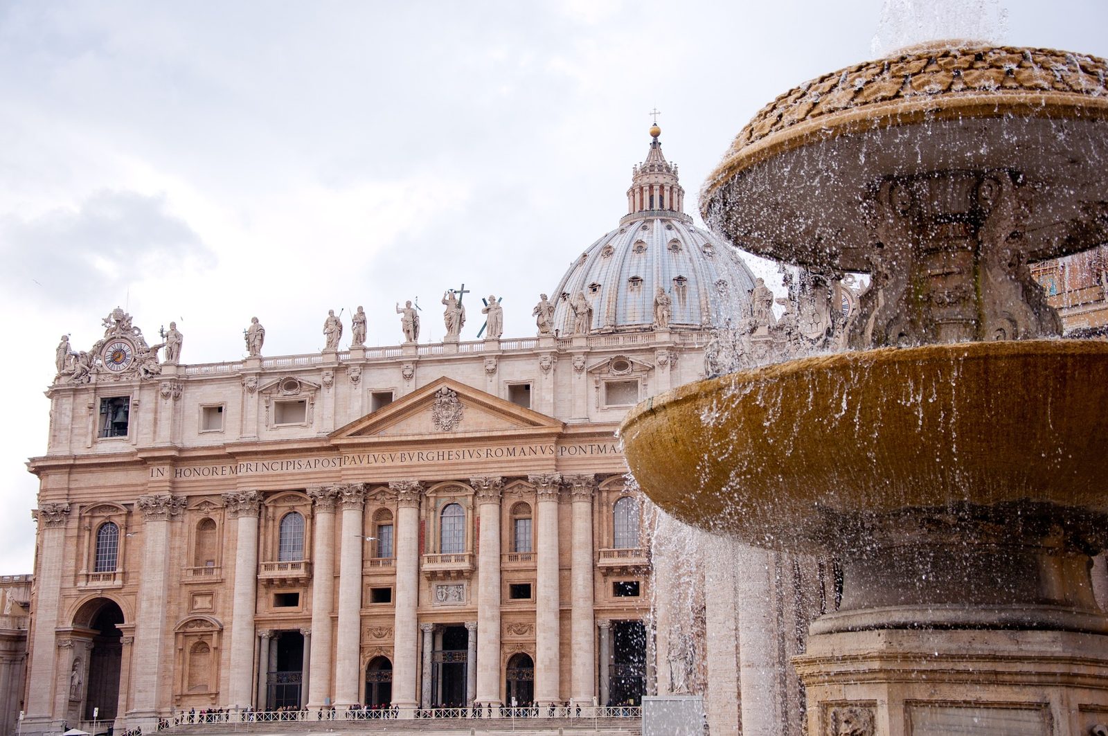The façade and dome of St Peter's Basilica, daytime, with one of the fountains of St Peter's Square in the foreground
